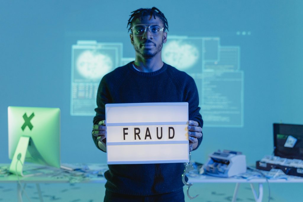 pexels-photo-6266500-6266500 Man holding a 'FRAUD' sign in a tech setting, symbolizing cybersecurity threats.